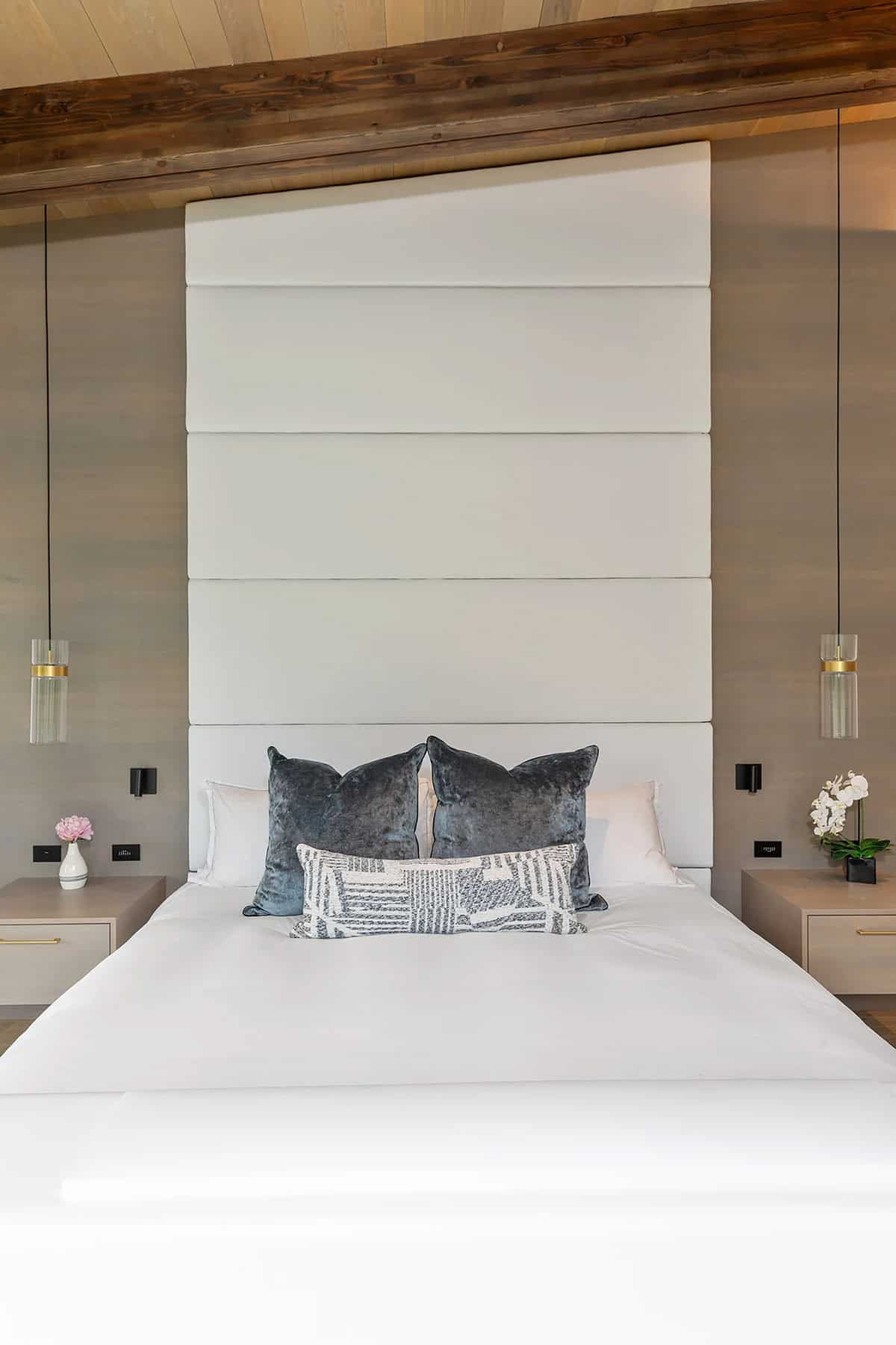 Primary bedroom with tall upholstered white headboard, velvet pillows, pendant sconces, and wood beam ceiling