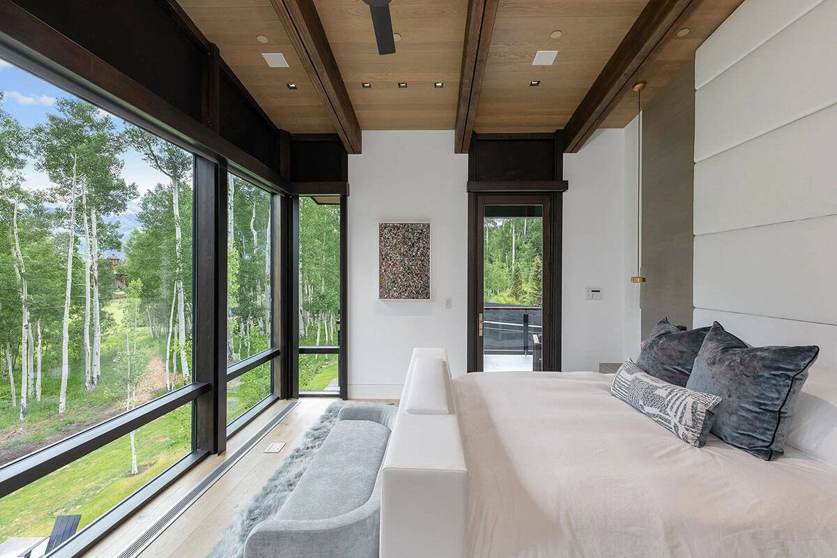 Primary bedroom with floor-to-ceiling windows, wood beam ceiling, upholstered bench, and aspen forest views