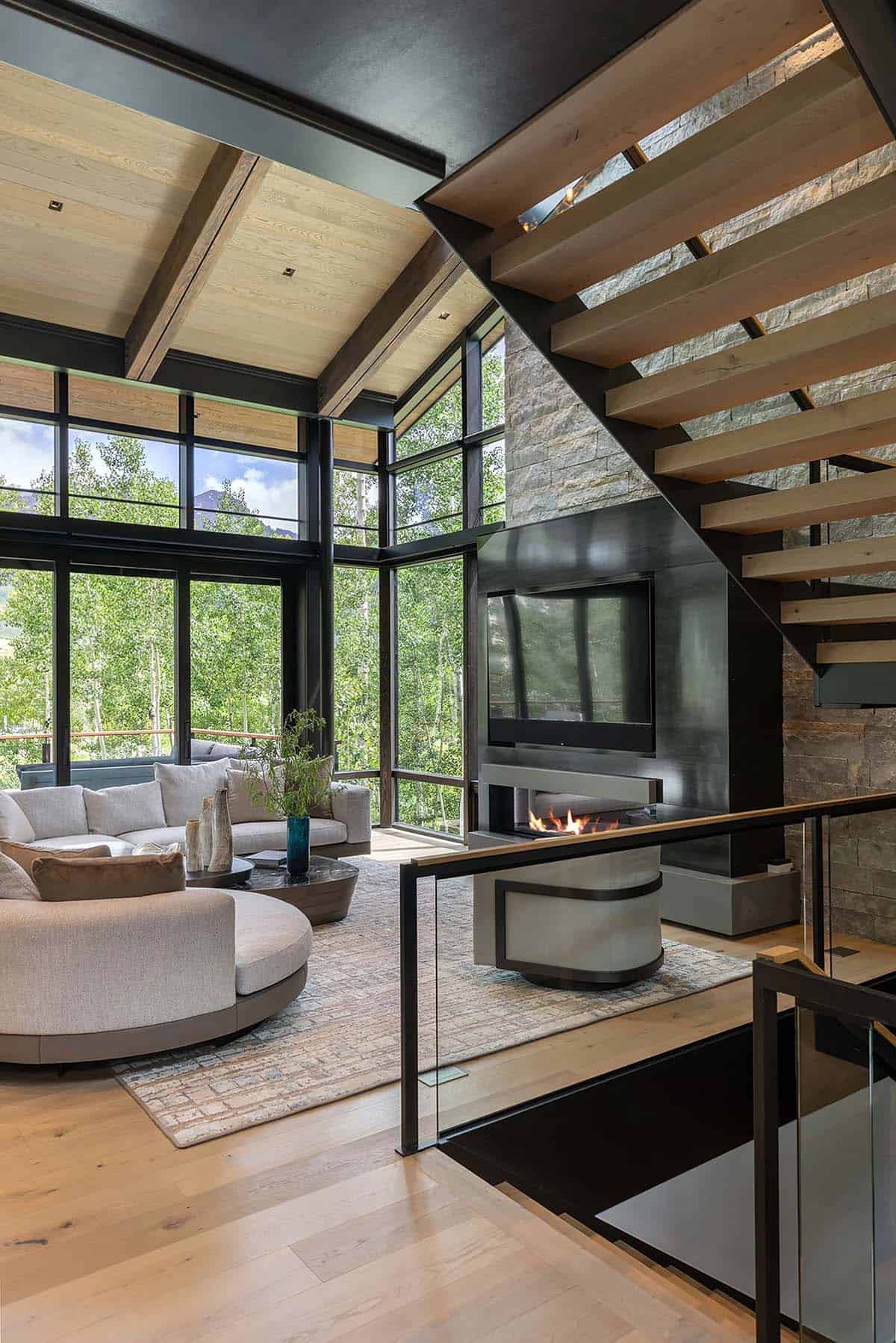 Living room view from staircase landing showing curved sectional, stone fireplace, and two-story glass walls