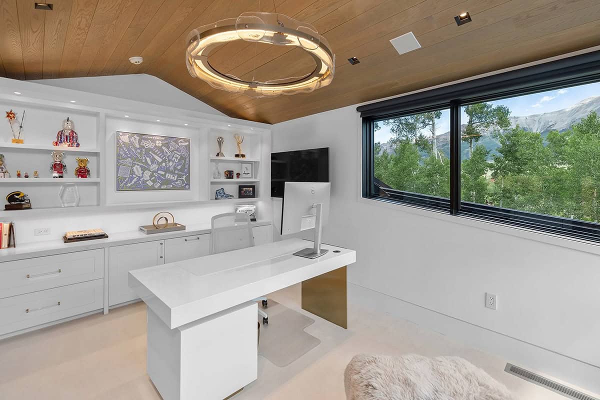 Home office with white floating desk, built-in shelving, circular pendant light, and mountain views through black-framed windows