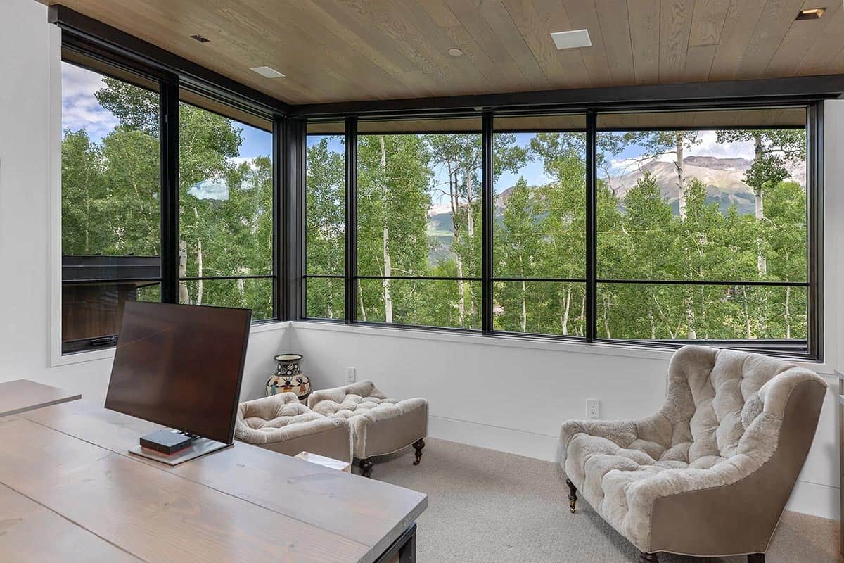 Home office or sitting room with tufted lounge chairs, wood desk, and panoramic corner windows with mountain and aspen views