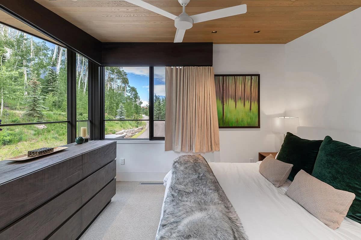 Guest bedroom with green velvet pillows, corner windows, dark dresser, and forest landscape artwork