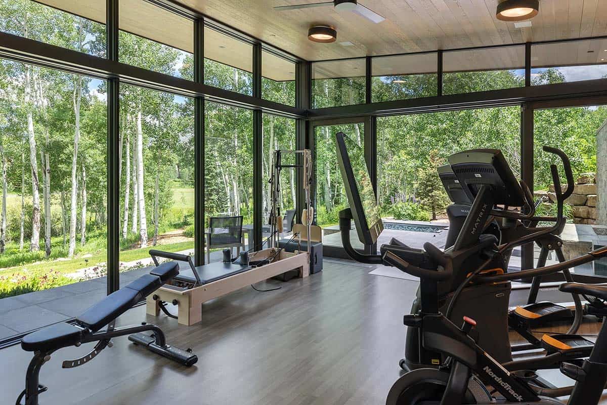 Glass-walled home gym with Pilates reformer, cardio equipment, wood ceiling, and wraparound aspen forest views