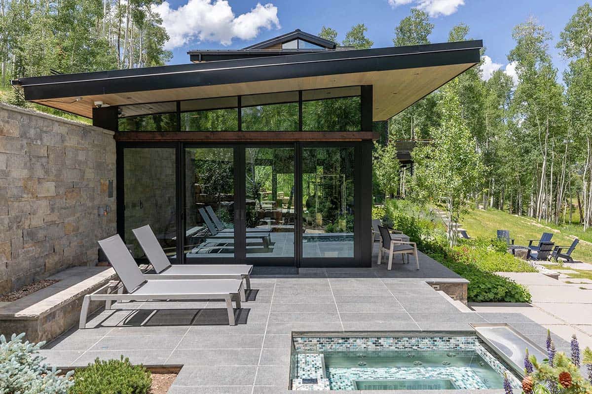 Exterior gym pavilion with cantilevered black roof, floor-to-ceiling glass, mosaic-tiled hot tub, and lounge chairs on slate patio