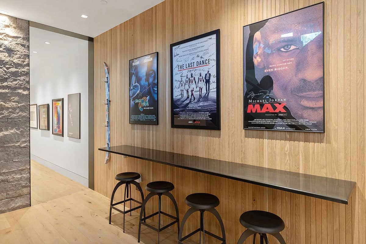 Lower-level sports bar hallway with framed basketball movie posters, long counter with black stools, and wood-slat wall