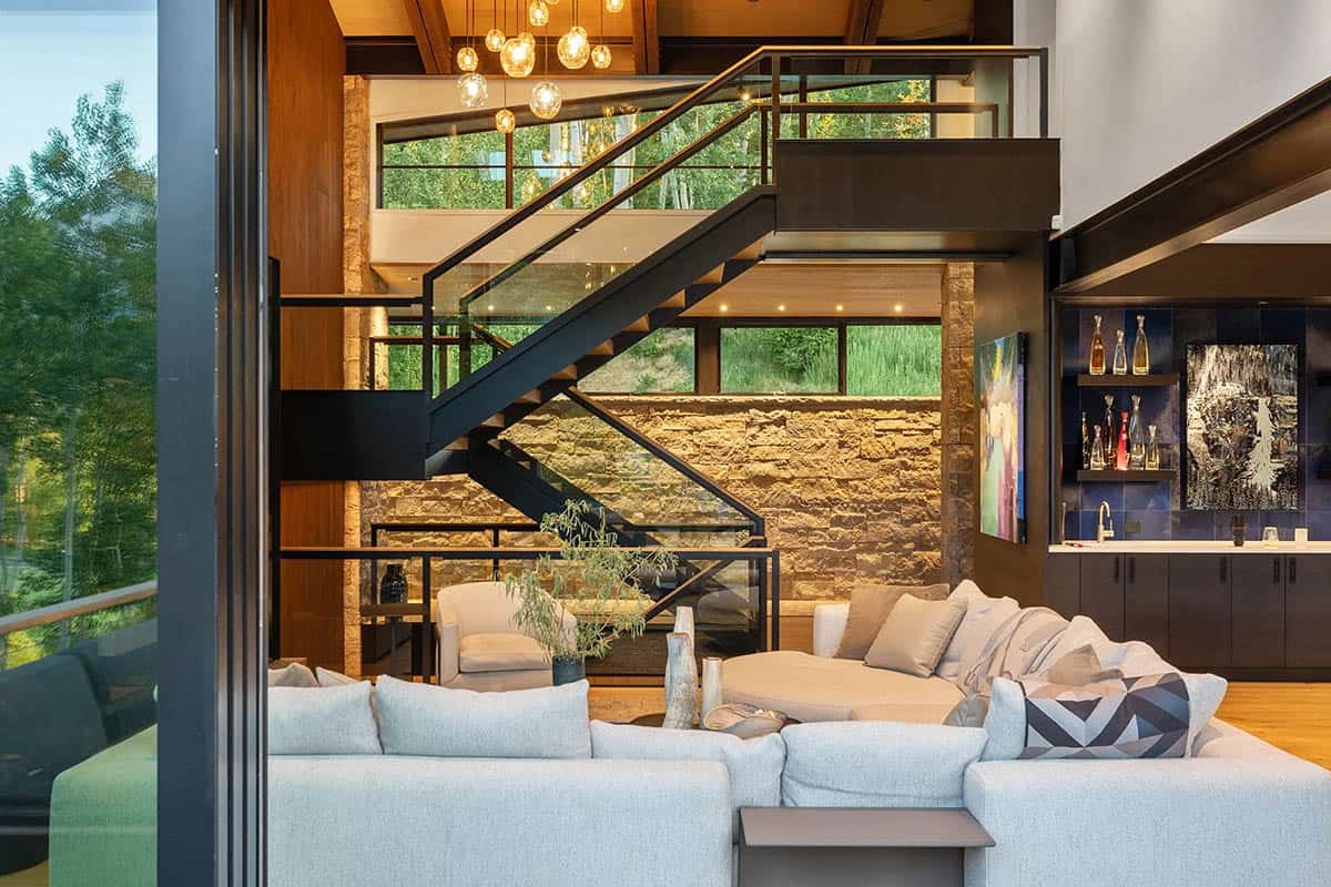 Evening interior view from deck showing living room with sectional sofa, floating staircase, and blue tile bar wall