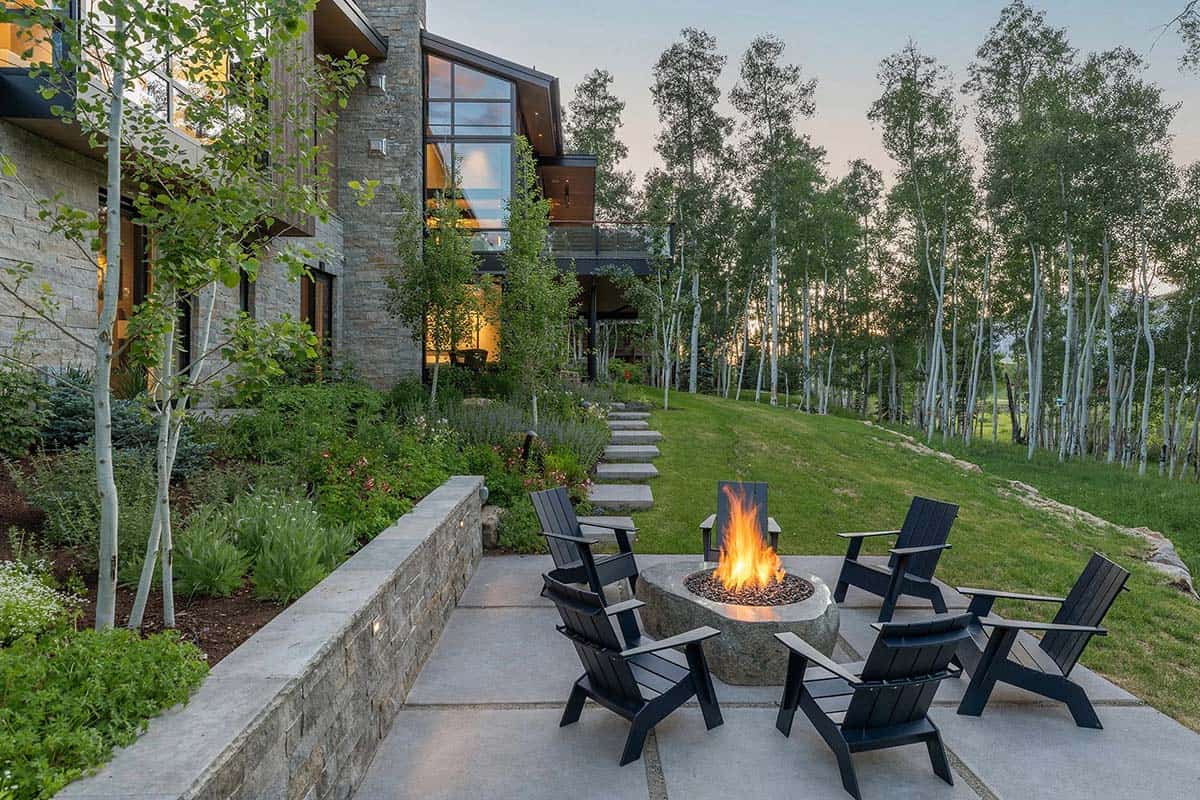 Dusk exterior with Adirondack chairs around lit stone fire bowl, terraced garden, and glowing mountain home facade