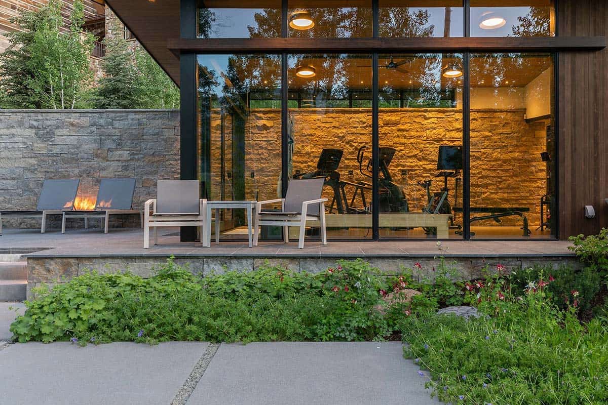 Dusk exterior of glass gym pavilion with lit interior, outdoor lounge chairs, plunge pool flame, and flowering garden