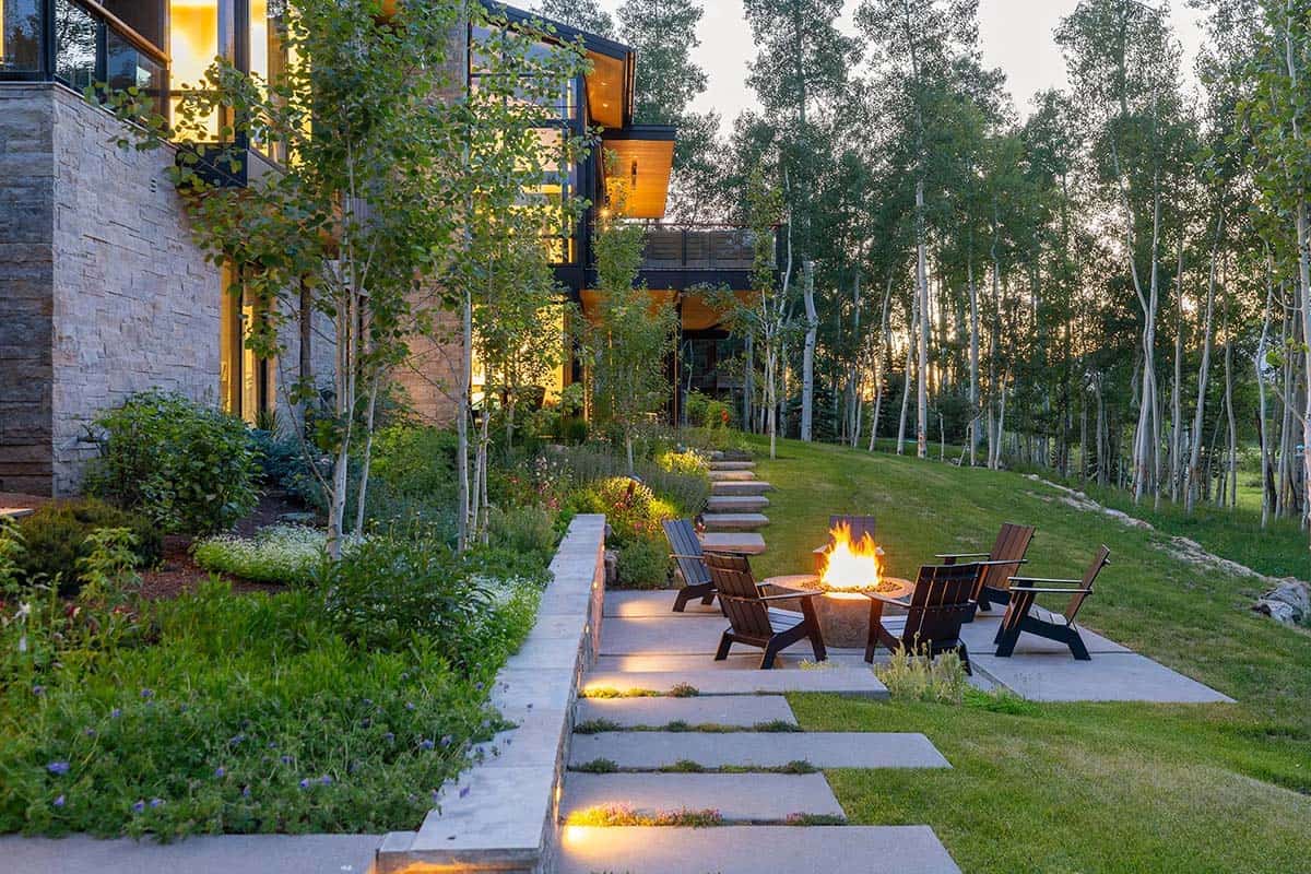 Dusk landscaping view with lit flagstone steps, fire pit seating, lush perennial borders, and glowing home exterior