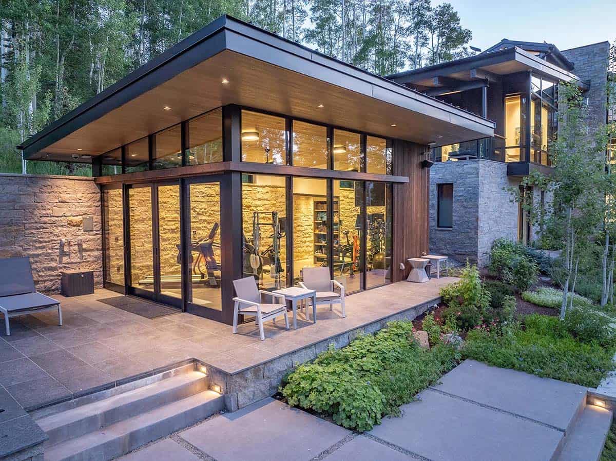 Dusk exterior of gym wing with cantilevered black roof, floor-to-ceiling glass walls, patio seating, and landscaped grounds