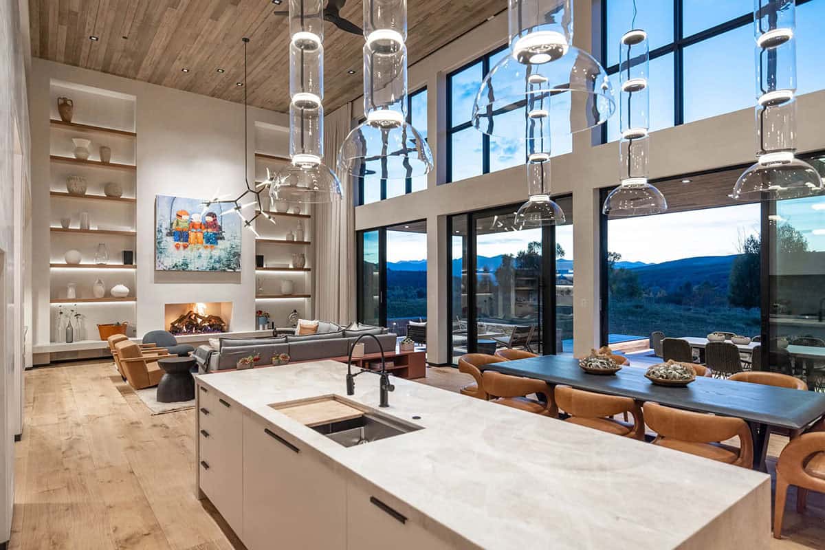 Great room overview showing kitchen island, dining table, living area with fireplace, and panoramic mountain views through steel-framed windows