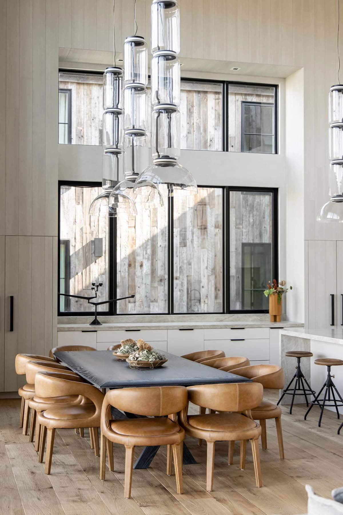 Dining area with dark slab table, curved cognac leather chairs, and cluster of glass pendant lights over double-height windows