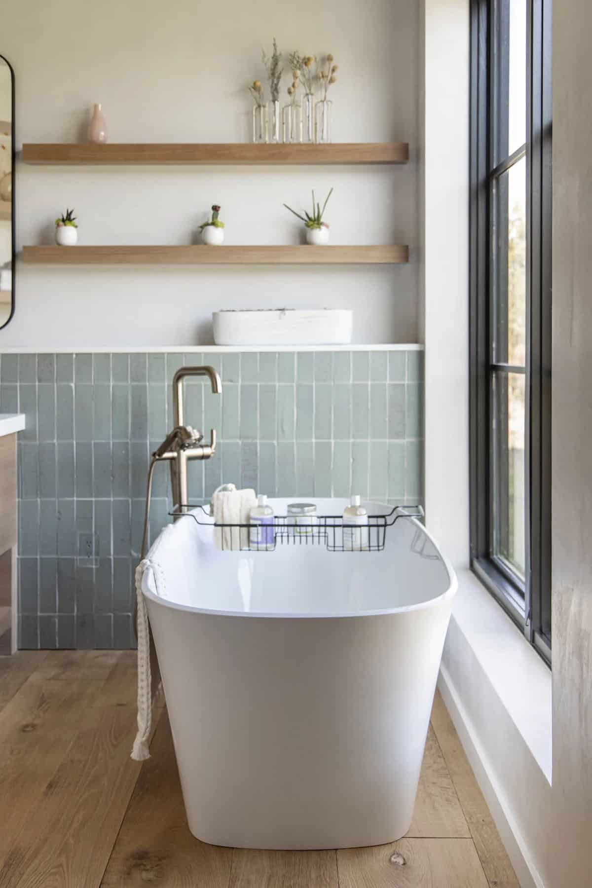 Spa-like bathroom with freestanding white soaking tub, sage green tile backsplash, and floating wood shelves