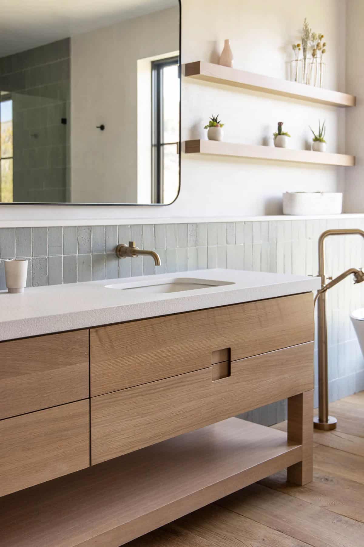 Bathroom vanity with white oak cabinetry, quartz countertop, wall-mounted brass faucet, and sage tile backsplash