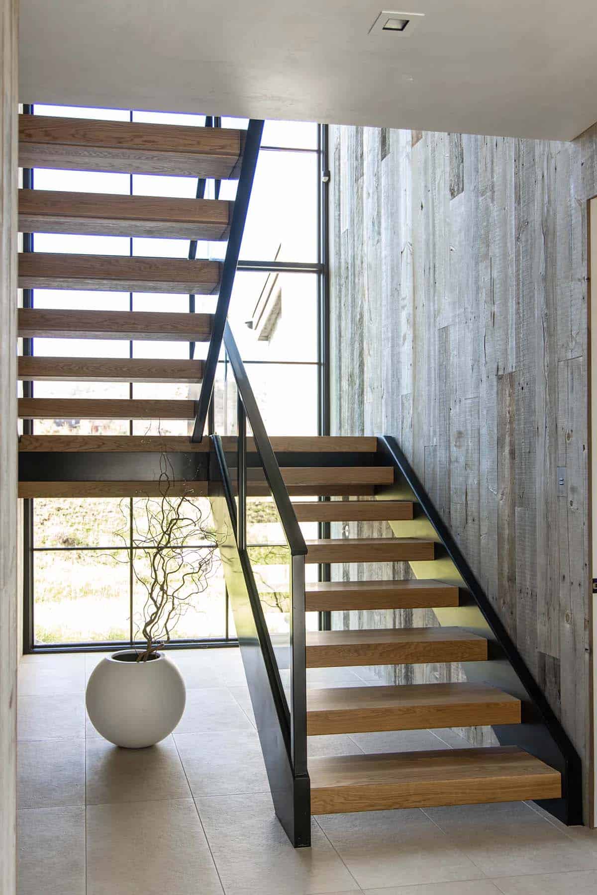 Open-riser oak staircase with black steel frame, reclaimed wood wall, and floor-to-ceiling windows flooding the entry with light