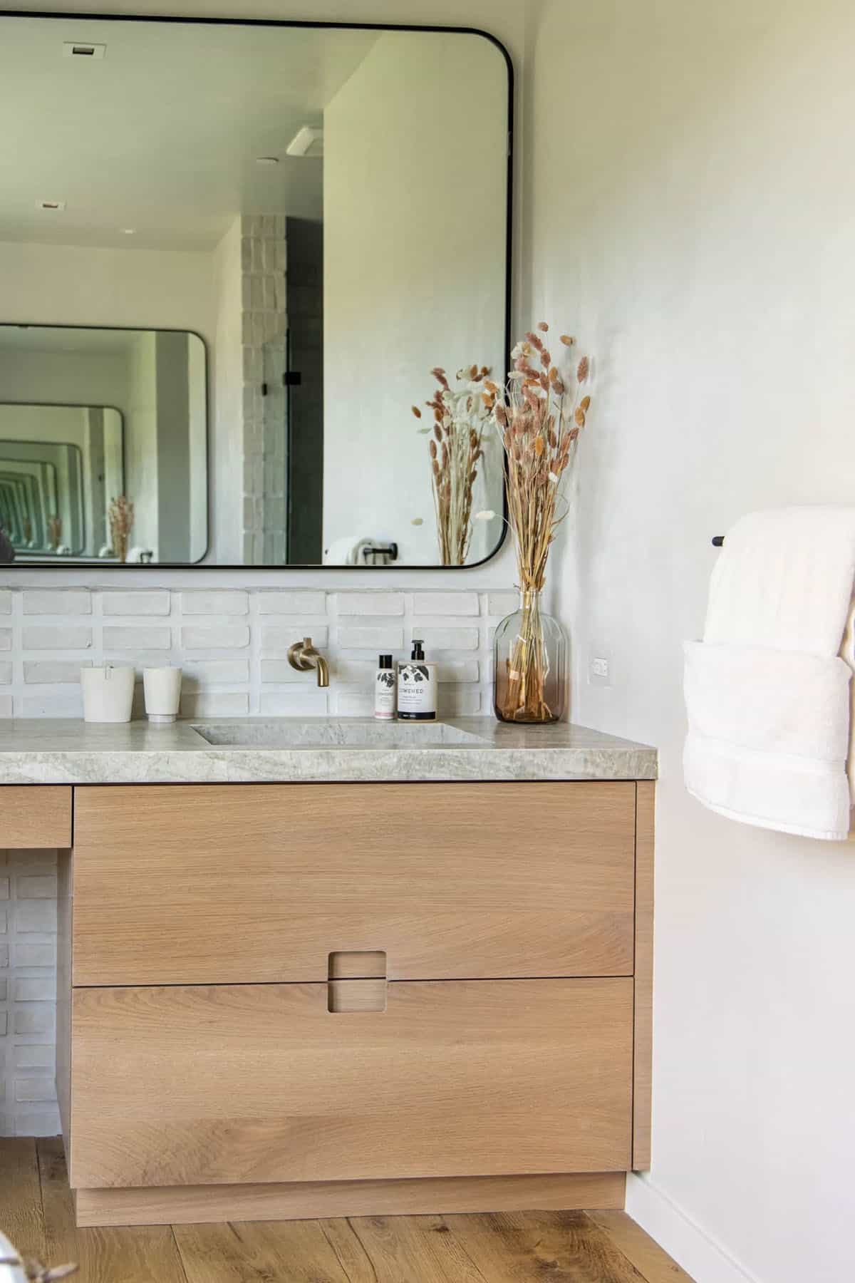 White oak bathroom vanity with stone countertop, white brick tile backsplash, and large black-framed mirror
