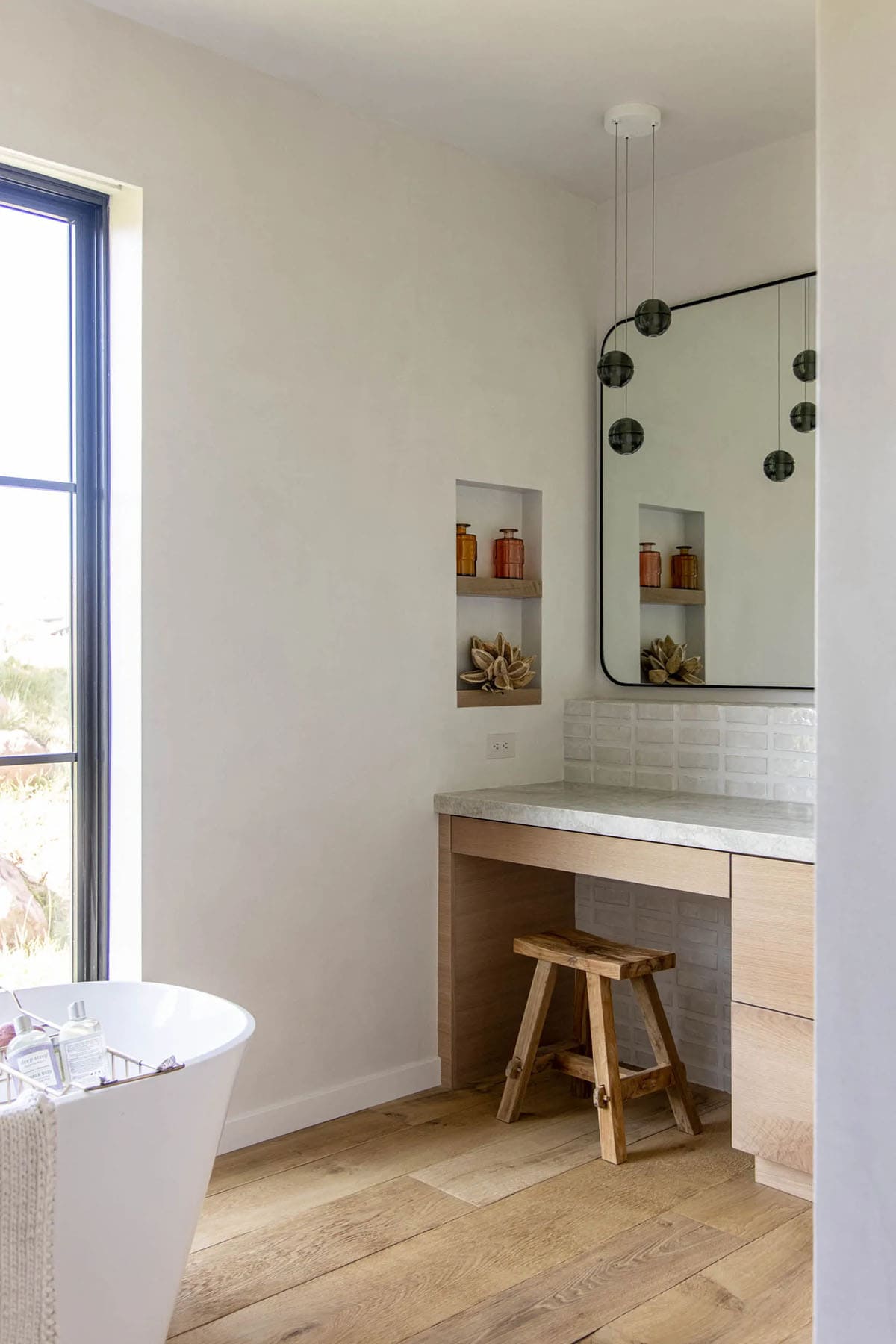 Bathroom dressing area with floating oak vanity, recessed wall niche, freestanding tub, and smoked glass globe pendant