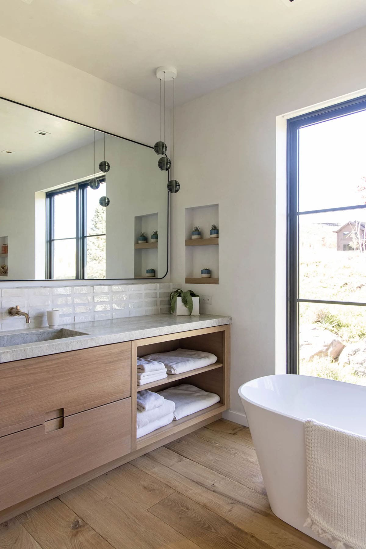 Primary bathroom with oak vanity, open towel shelving, freestanding soaking tub, and black-framed window overlooking landscape