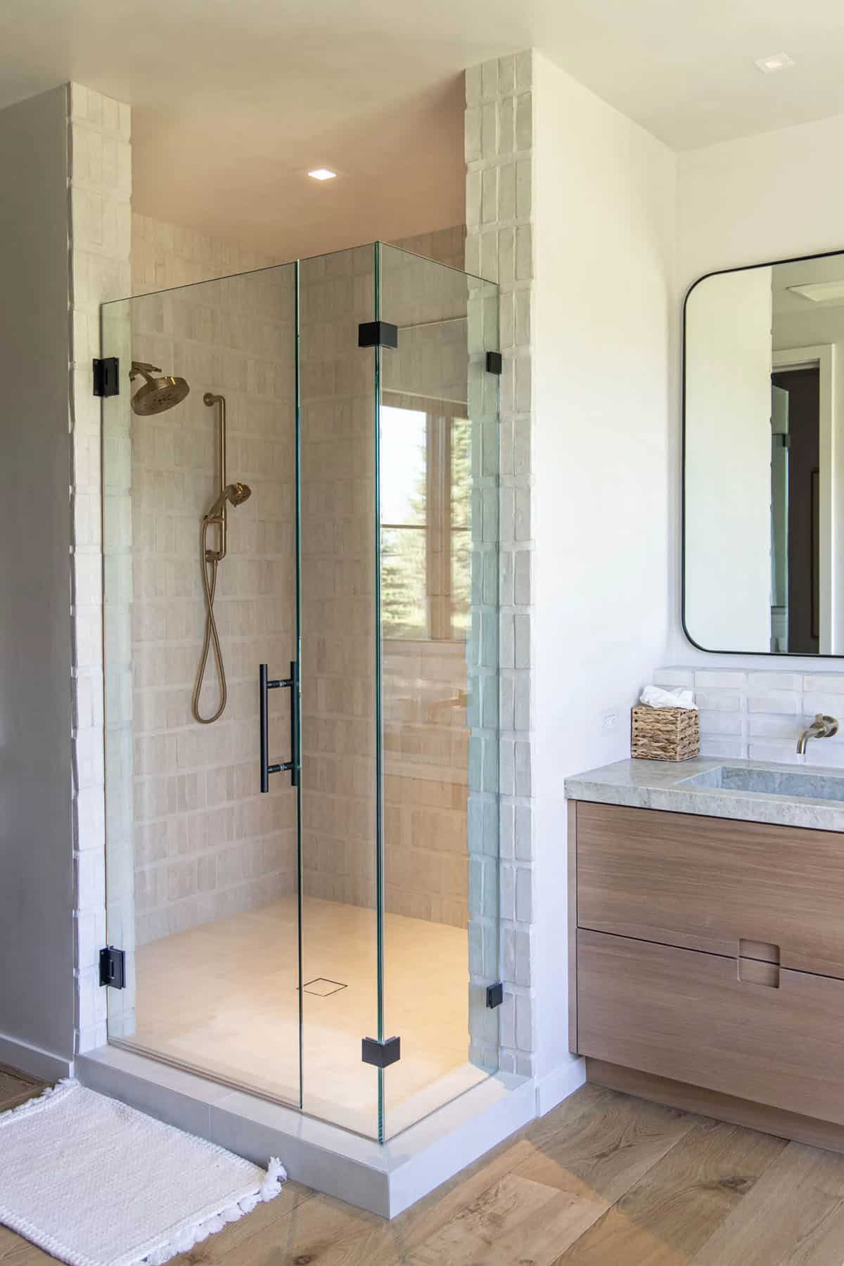 Glass-enclosed shower with cream zellige tile and brushed brass fixtures beside a floating oak vanity