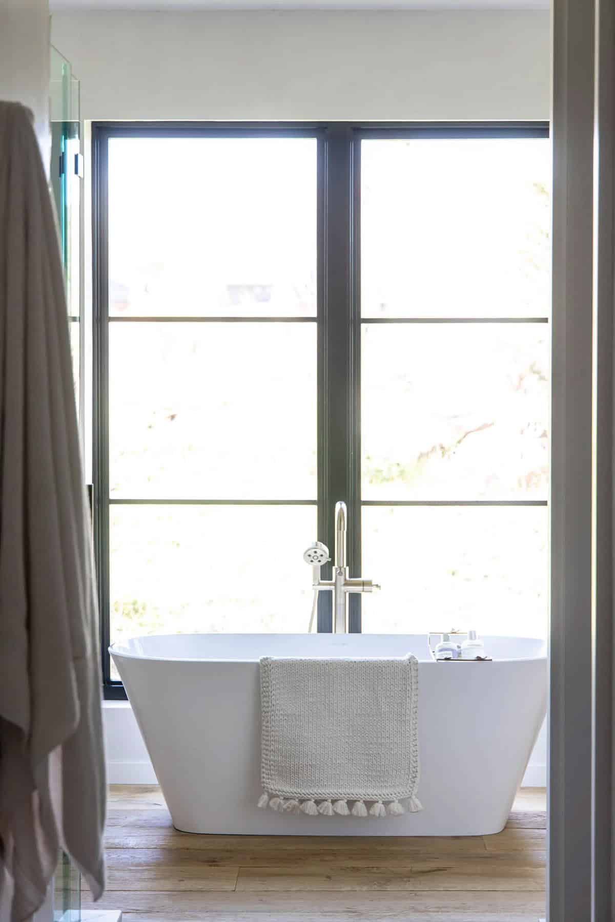 Freestanding white soaking tub centered before a large black steel-framed window with garden views