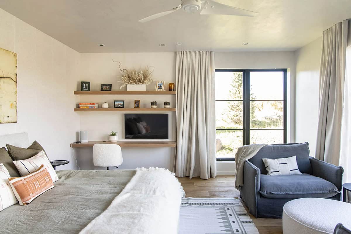 Primary bedroom sitting area with blue slipcovered chair, floating shelves, TV nook, and steel-framed window with tree views