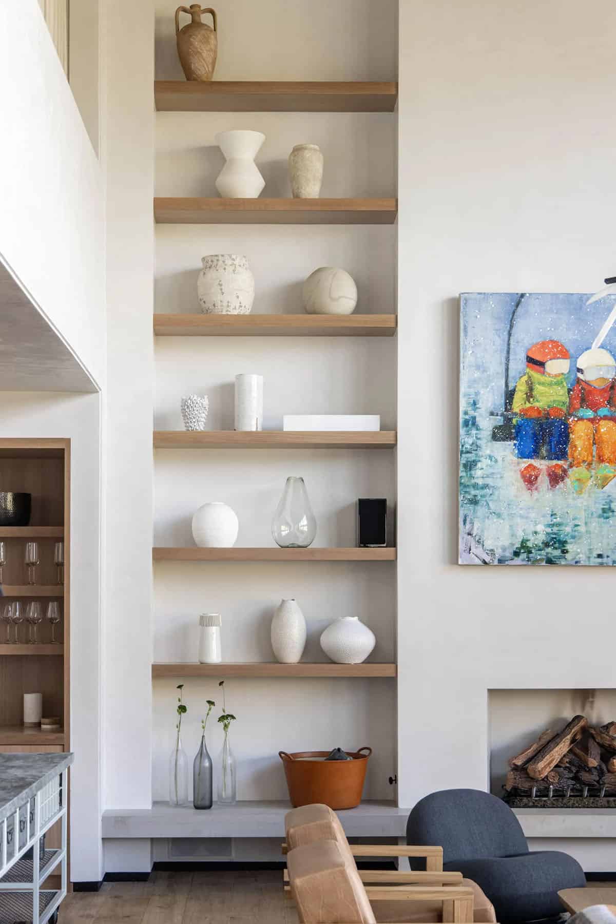 Tall built-in living room shelves styled with white ceramic vessels beside a lit fireplace and colorful ski painting