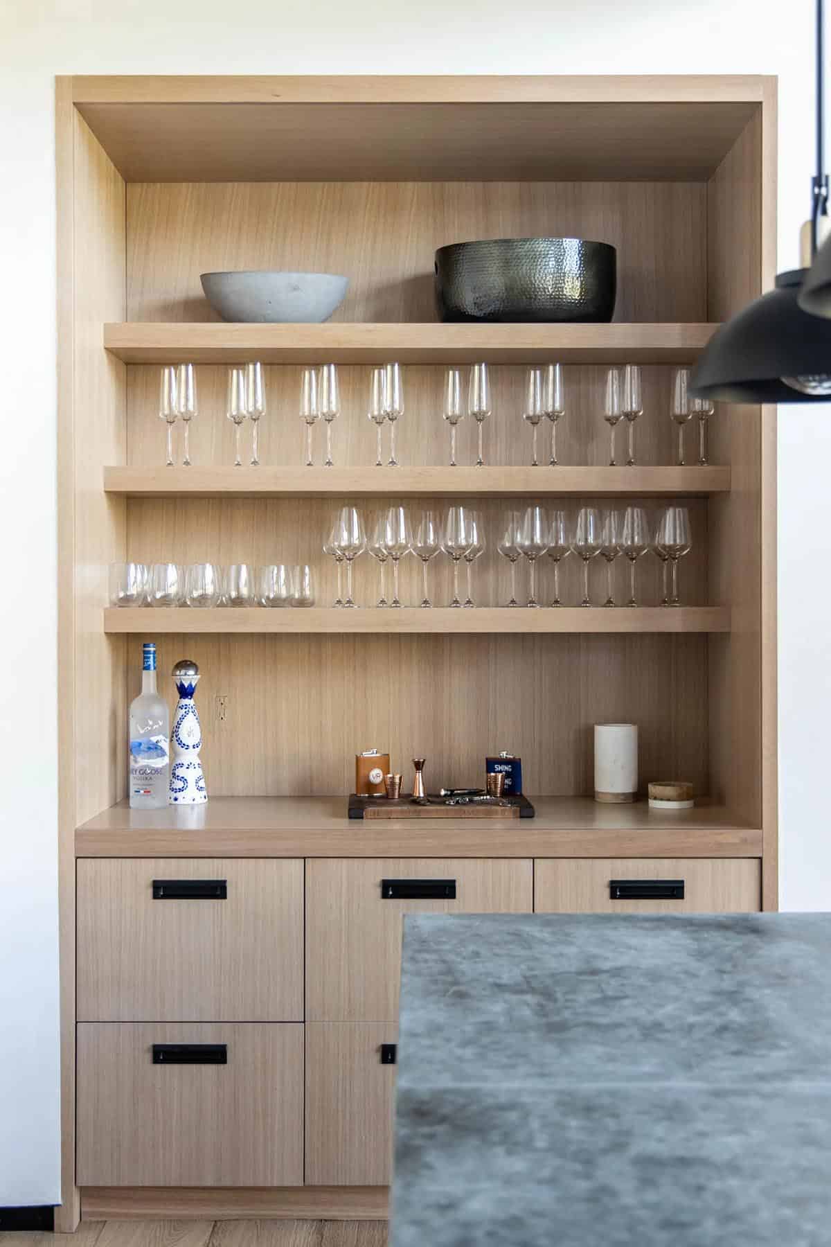 Close-up of white oak built-in bar with rows of stemware, spirit bottles, and matte black drawer pulls