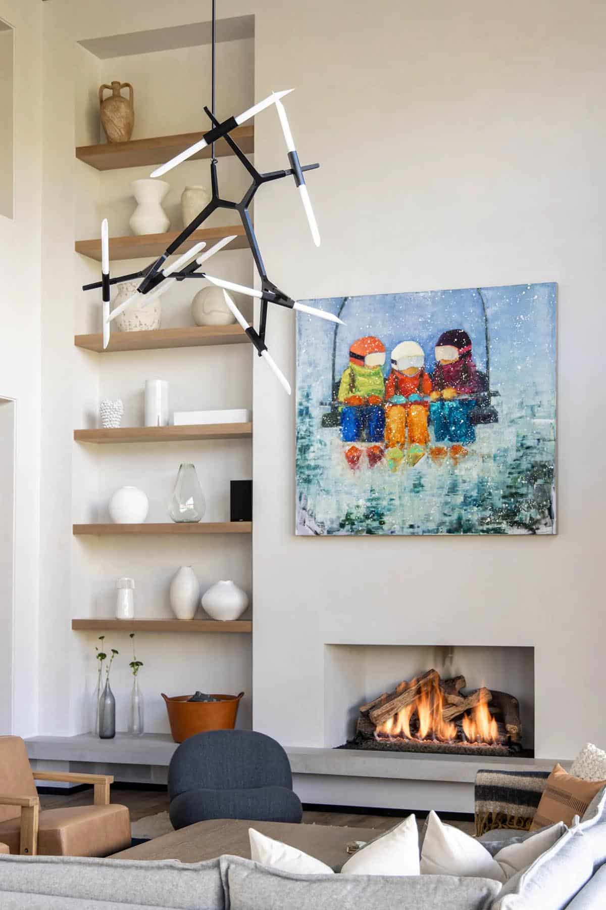 Living room fireplace wall with branching black chandelier, floor-to-ceiling oak shelves, and colorful ski lift painting