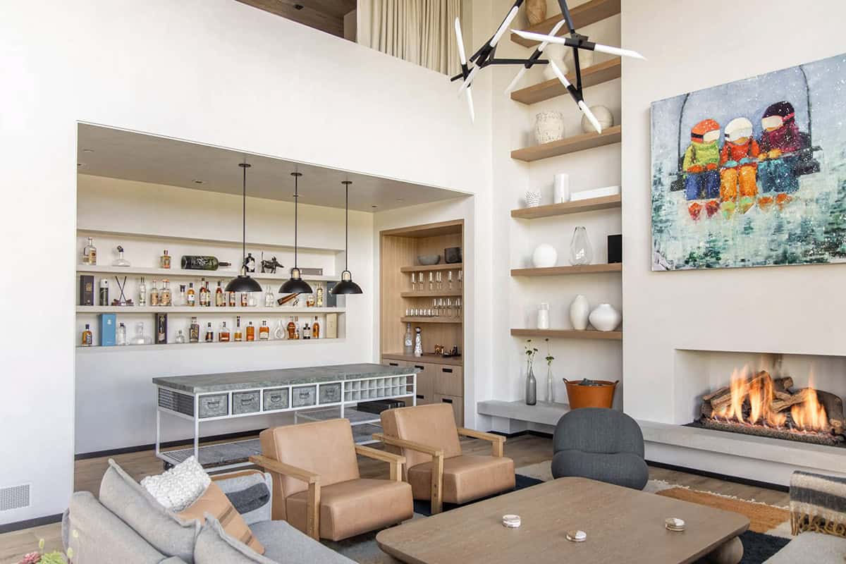 Open-plan living area showing bar niche, fireplace, leather armchairs, and colorful ski painting with branching chandelier 