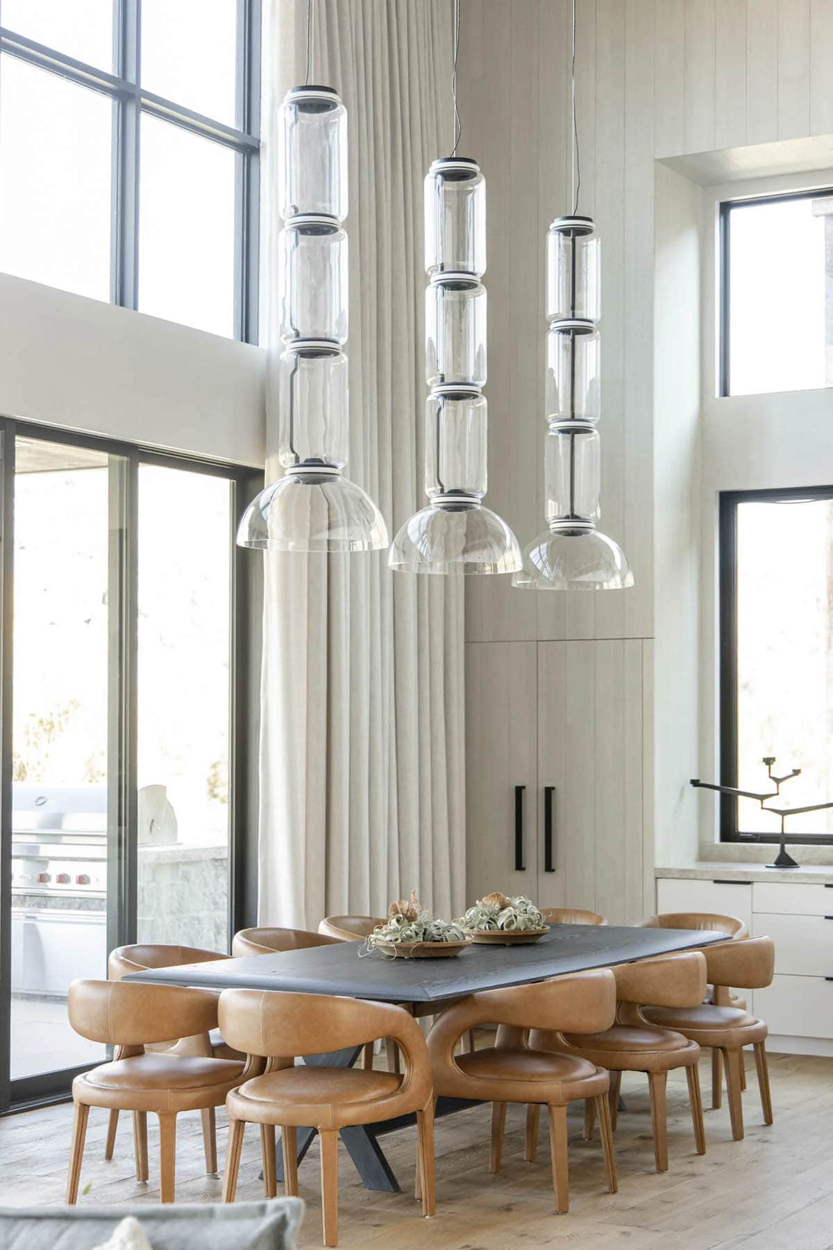 Dining area with dark slab table, cognac leather chairs, and trio of stacked glass cylinder pendants against tall windows