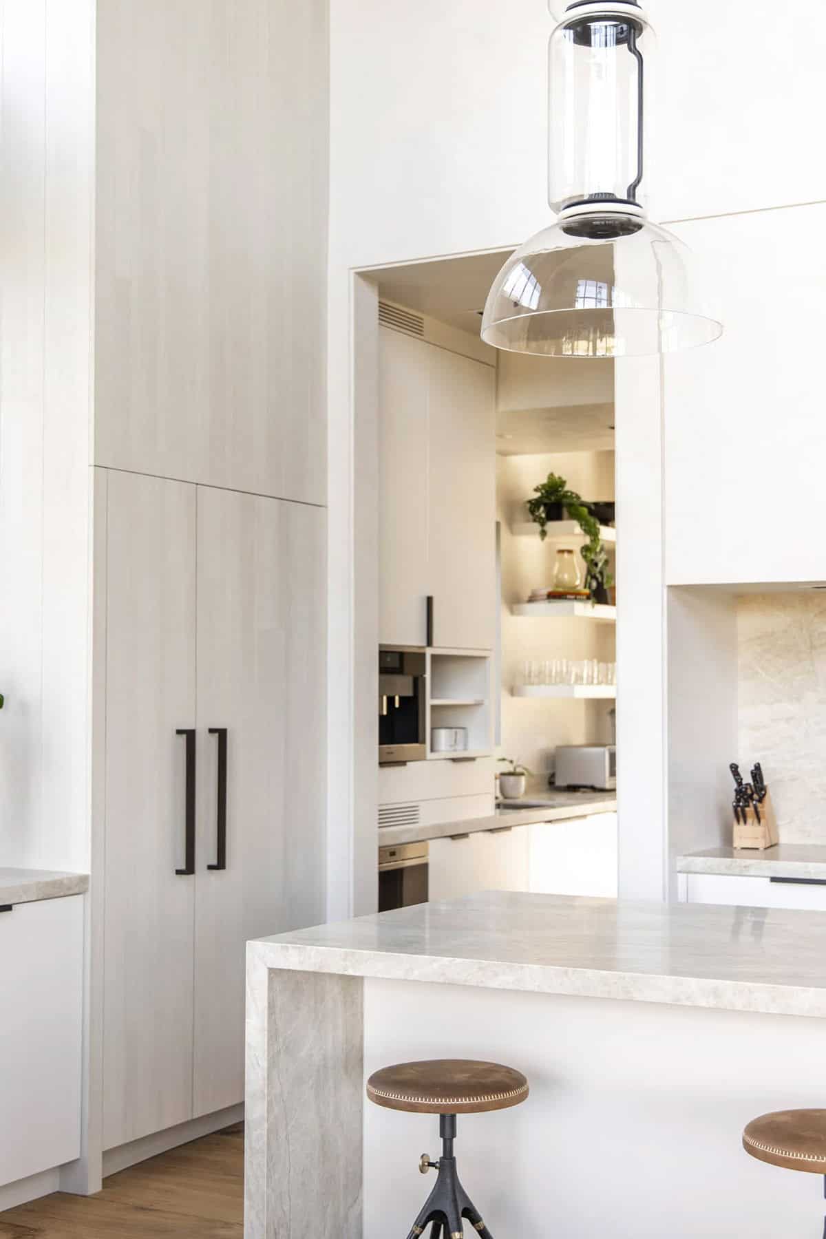 Kitchen island with travertine waterfall edge, industrial stools, and view through to illuminated scullery