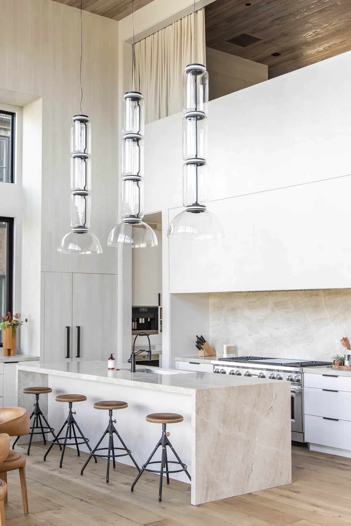 White kitchen with travertine island, industrial bar stools, stacked glass pendants, and pro range with stone backsplash