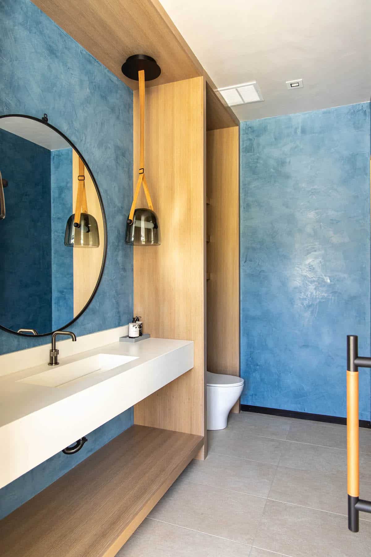 Bold blue venetian plaster powder room with floating white vanity, round black mirror, and leather-strap smoked glass pendants