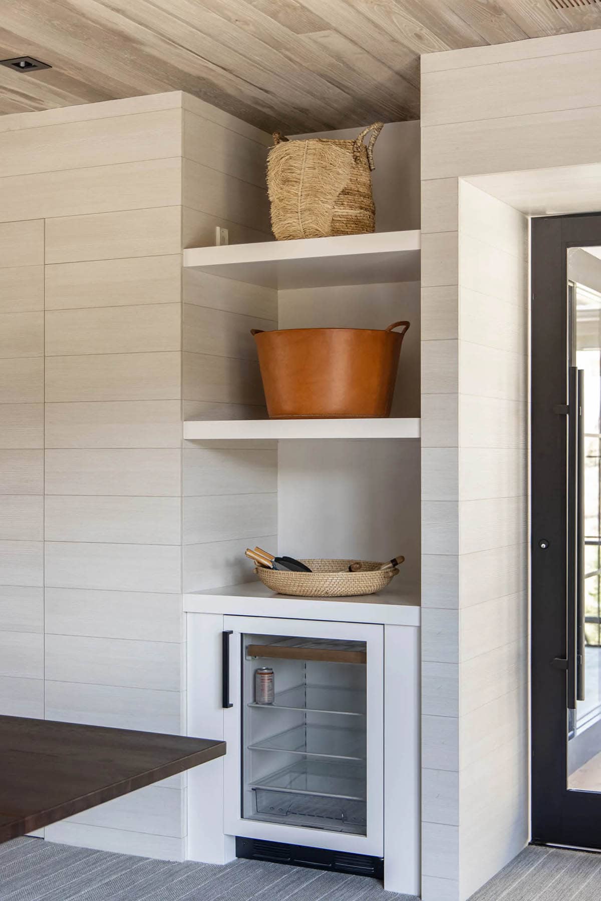 Lower-level game room corner with built-in shelves, glass-door beverage fridge, woven baskets, and leather bucket