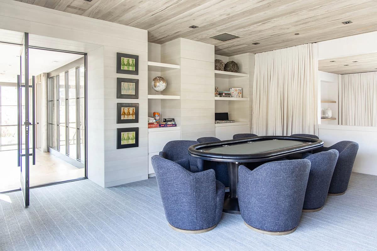 Game room with oval poker table, upholstered blue tub chairs, built-in shelving, and wood plank ceiling