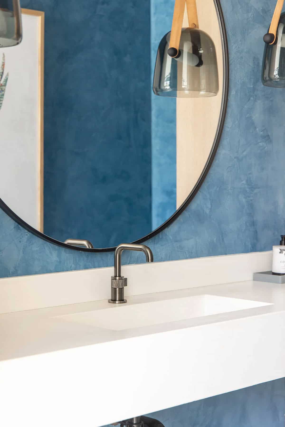 Close-up of powder room round mirror, brushed steel faucet, white integrated sink, and blue plaster wall