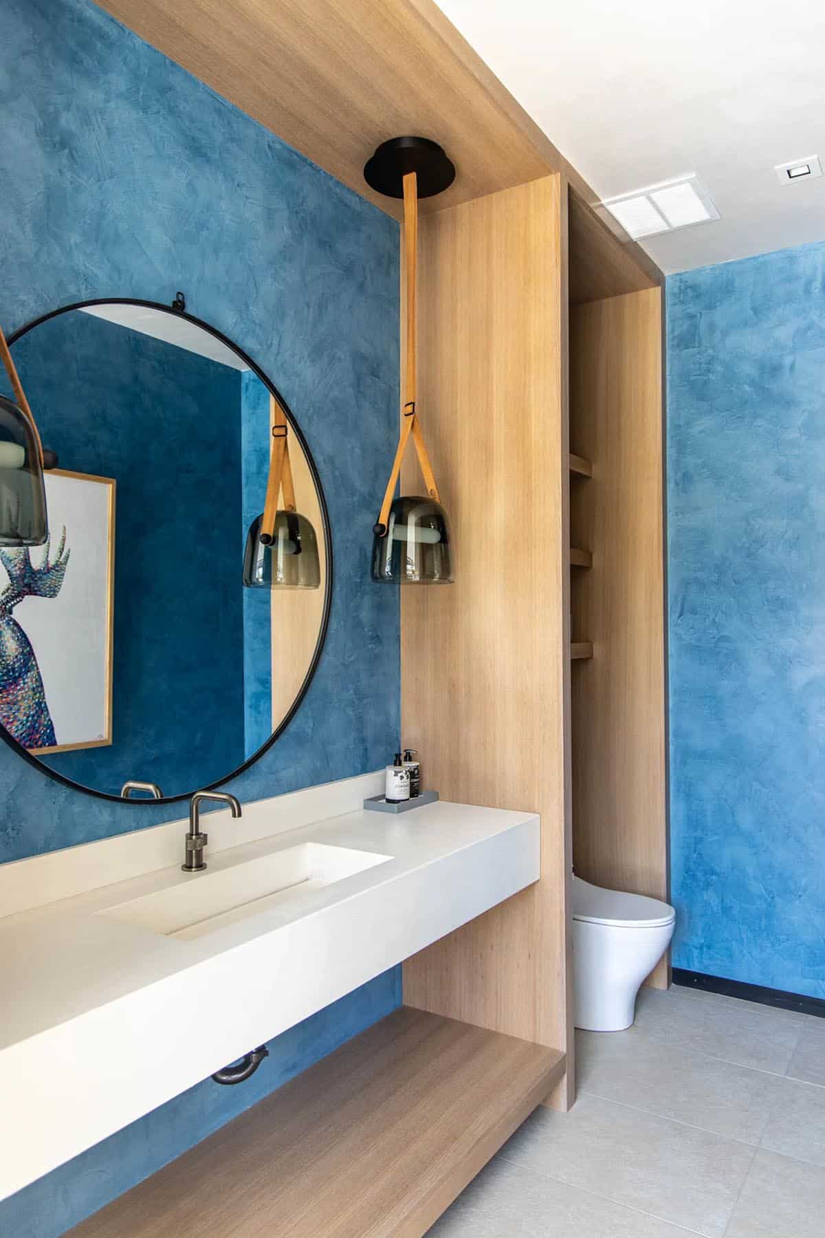 Blue plaster bathroom with floating white vanity, round mirror, smoked glass pendants on leather straps, and wall-hung toilet