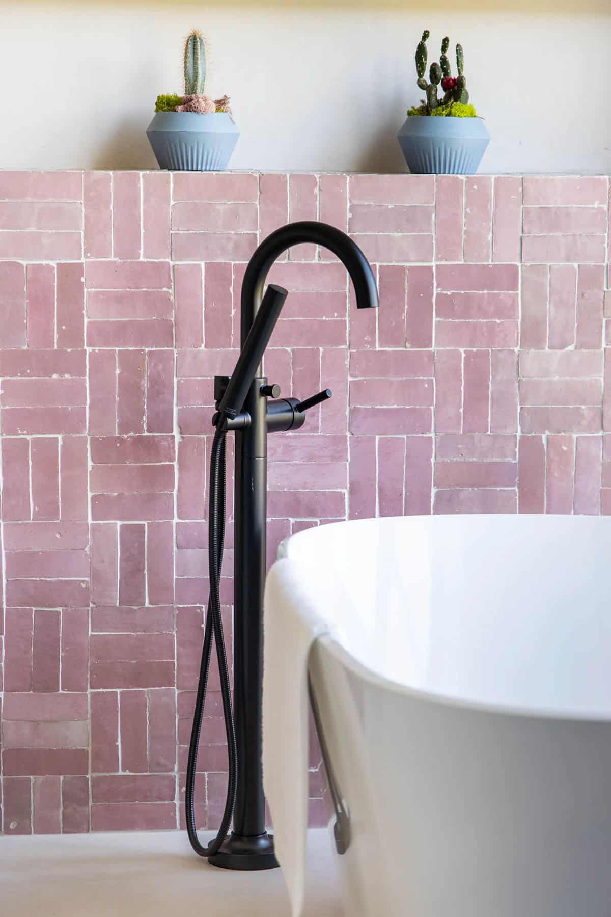 Matte black freestanding tub filler against pink zellige tile backsplash with potted cacti on ledge