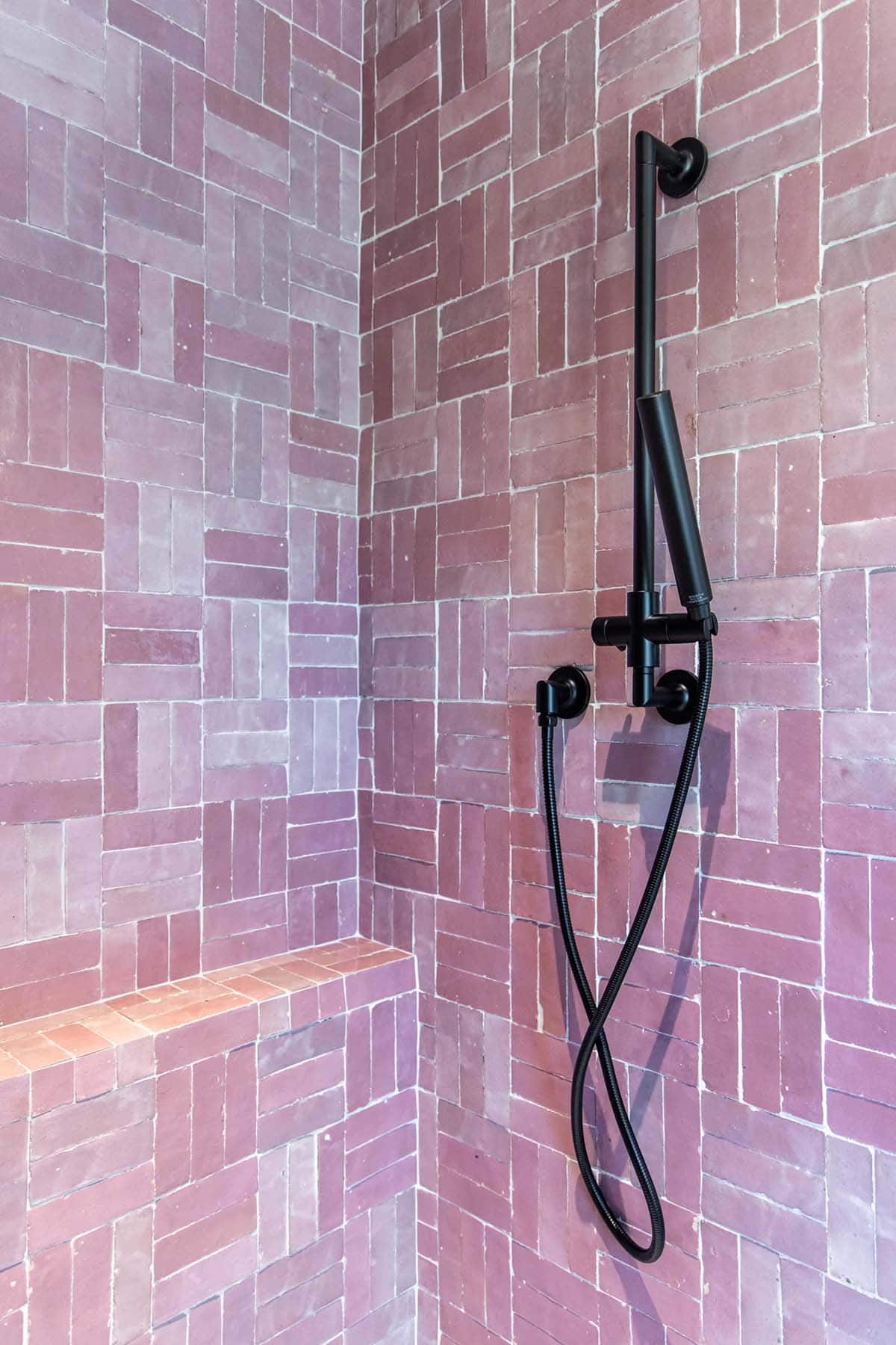 Pink zellige tile walk-in shower corner with black handheld shower on adjustable rail and built-in bench