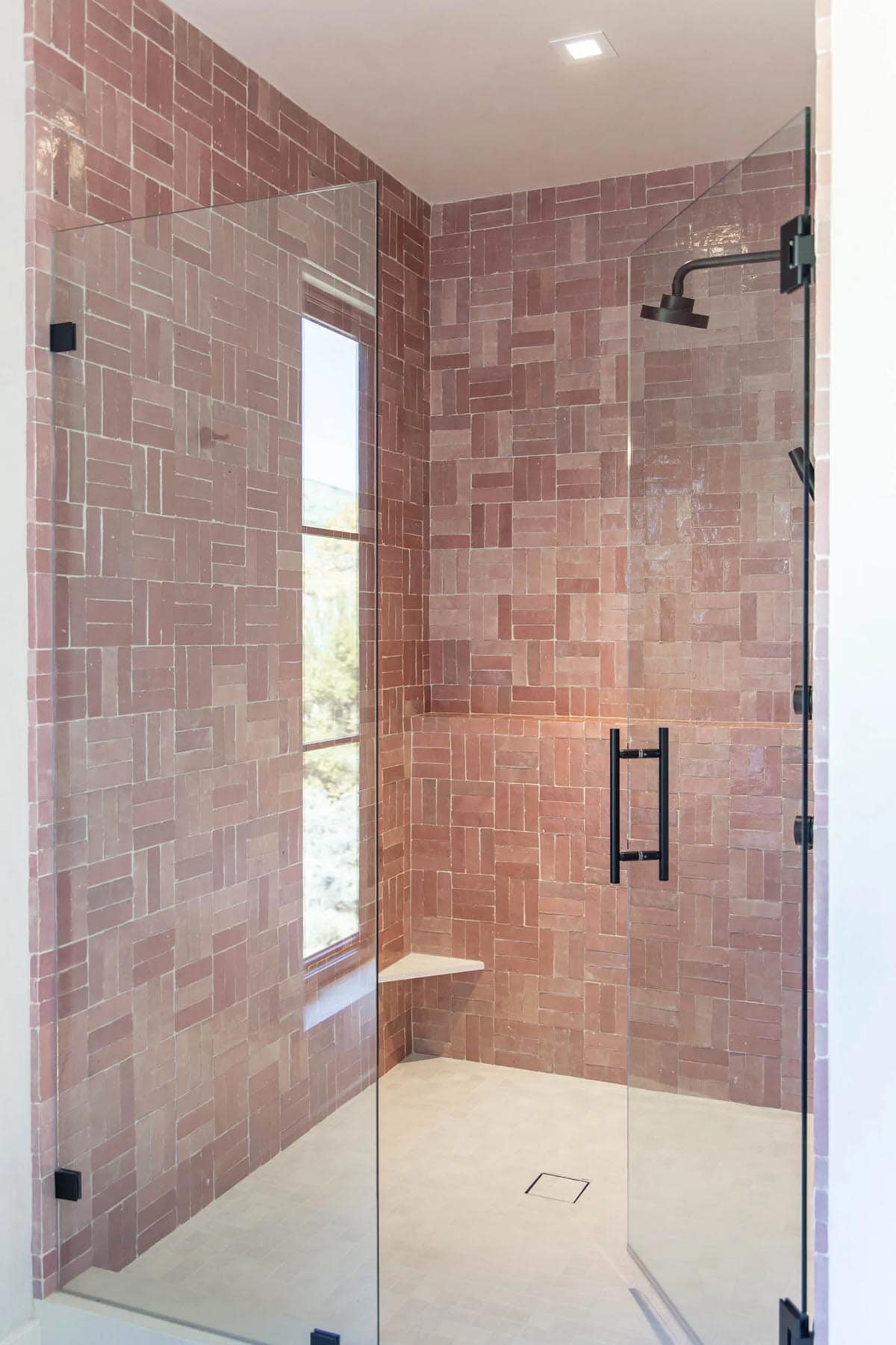 Glass-enclosed shower with floor-to-ceiling pink zellige tile in mixed herringbone pattern, black rainfall head, and corner shelf