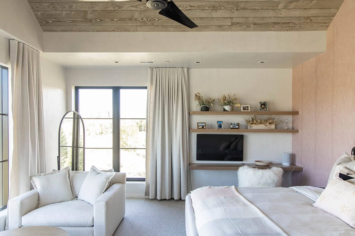 Bedroom sitting area with white slipcovered chair, linen drapes, floating oak shelves, TV nook, and mountain views