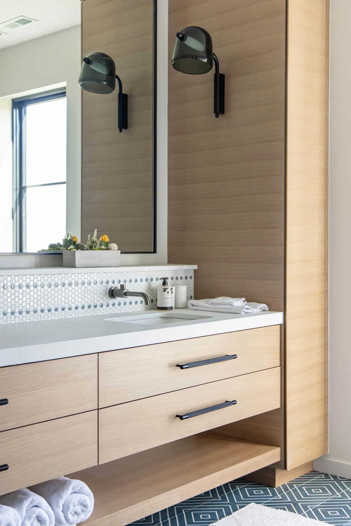 Close-up of white oak bathroom vanity with smoked glass sconce, penny tile backsplash, and black matte hardware