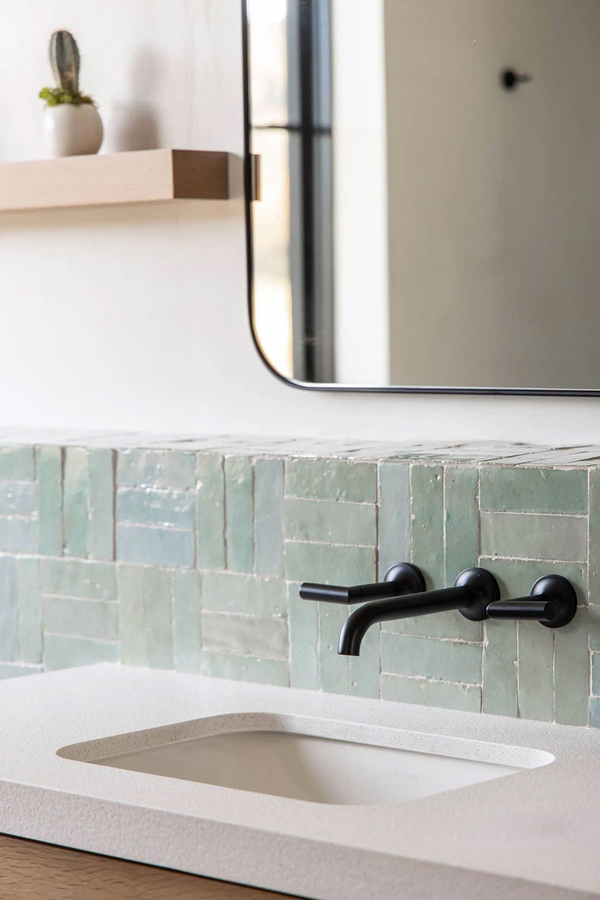 Close-up of white quartz bathroom sink with matte black wall-mounted faucet and aqua zellige tile backsplash