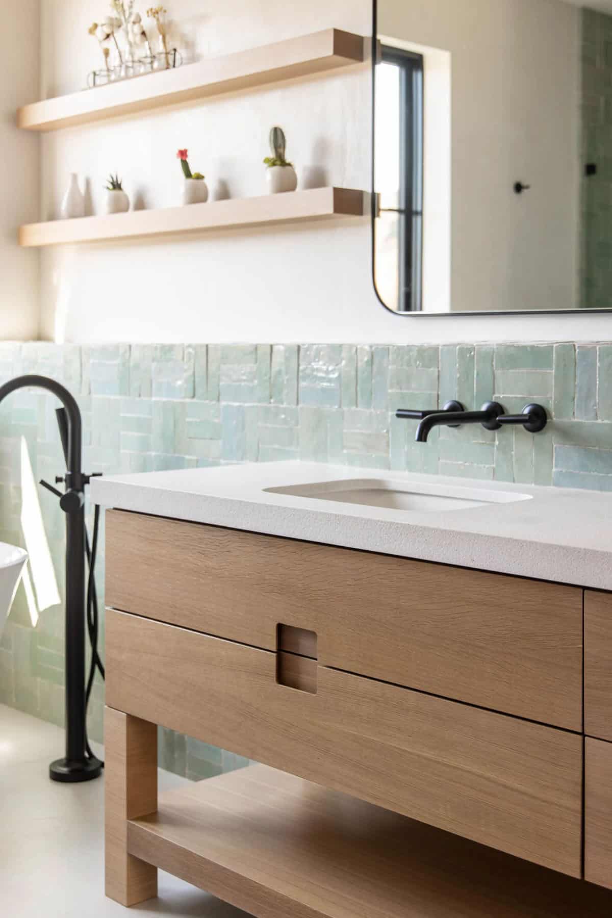 White oak bathroom vanity with quartz top, aqua zellige backsplash, floating oak shelves with cacti, and black freestanding tub filler