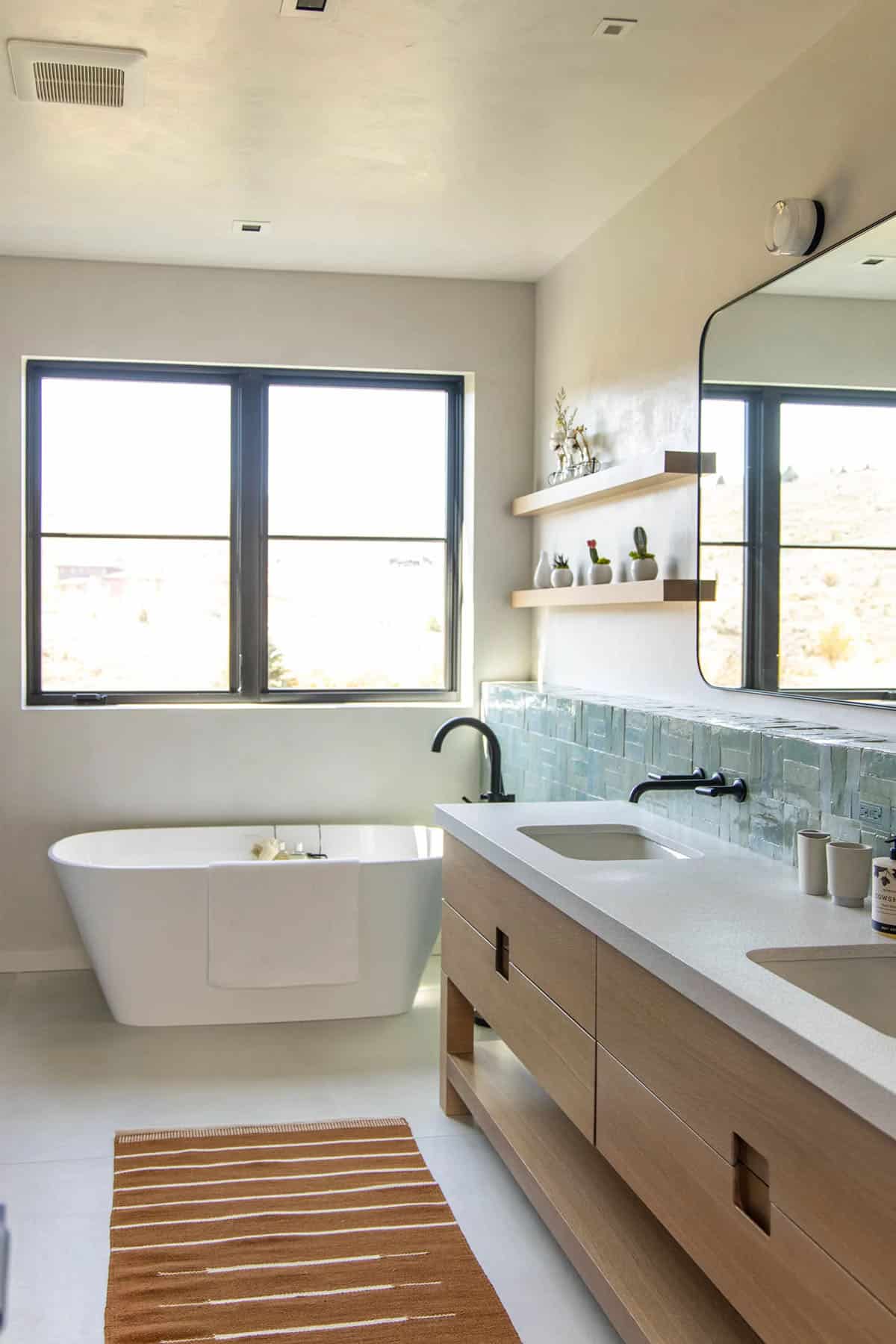 Bright bathroom with white oak vanity, aqua zellige backsplash, freestanding soaking tub, and mountain views through black-framed windows