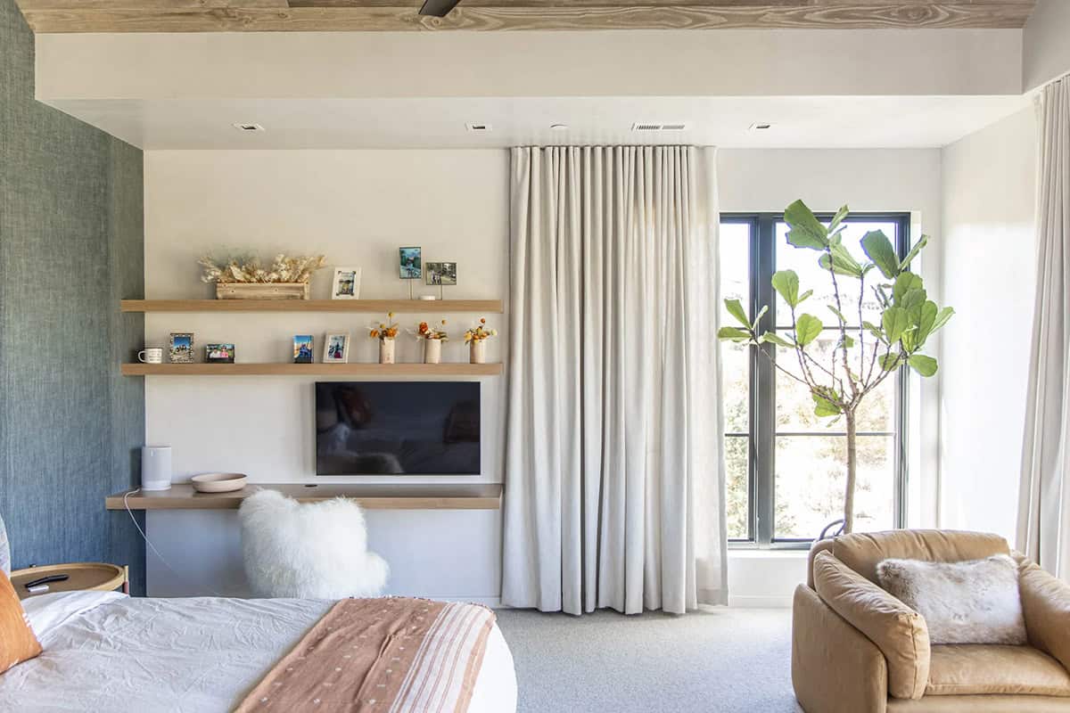 Bedroom media wall with floating oak shelves, TV nook, linen drapes, caramel armchair, and fiddle leaf fig