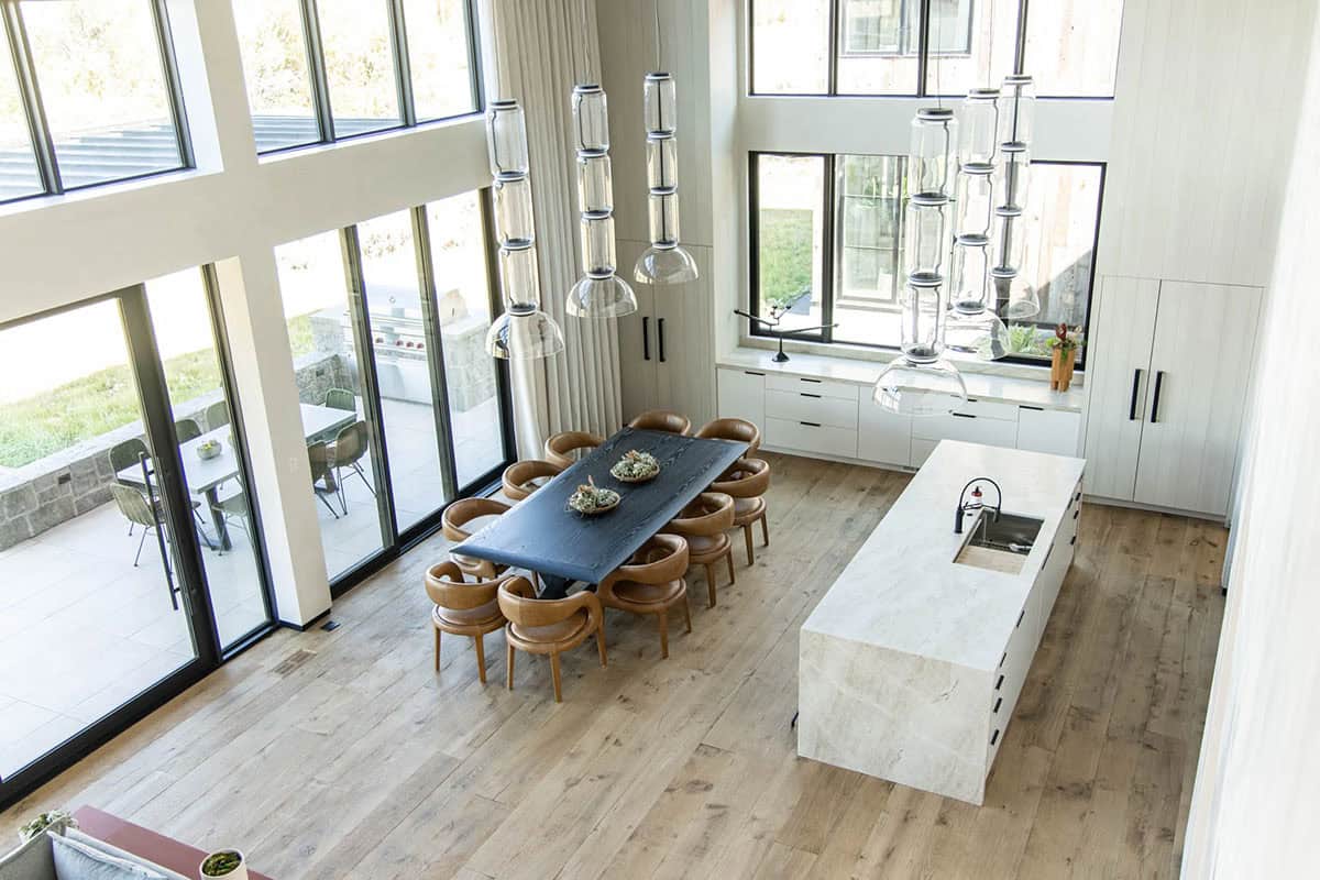 Aerial view of open-plan kitchen and dining with travertine island, dark slab dining table, leather chairs, and stacked glass pendants