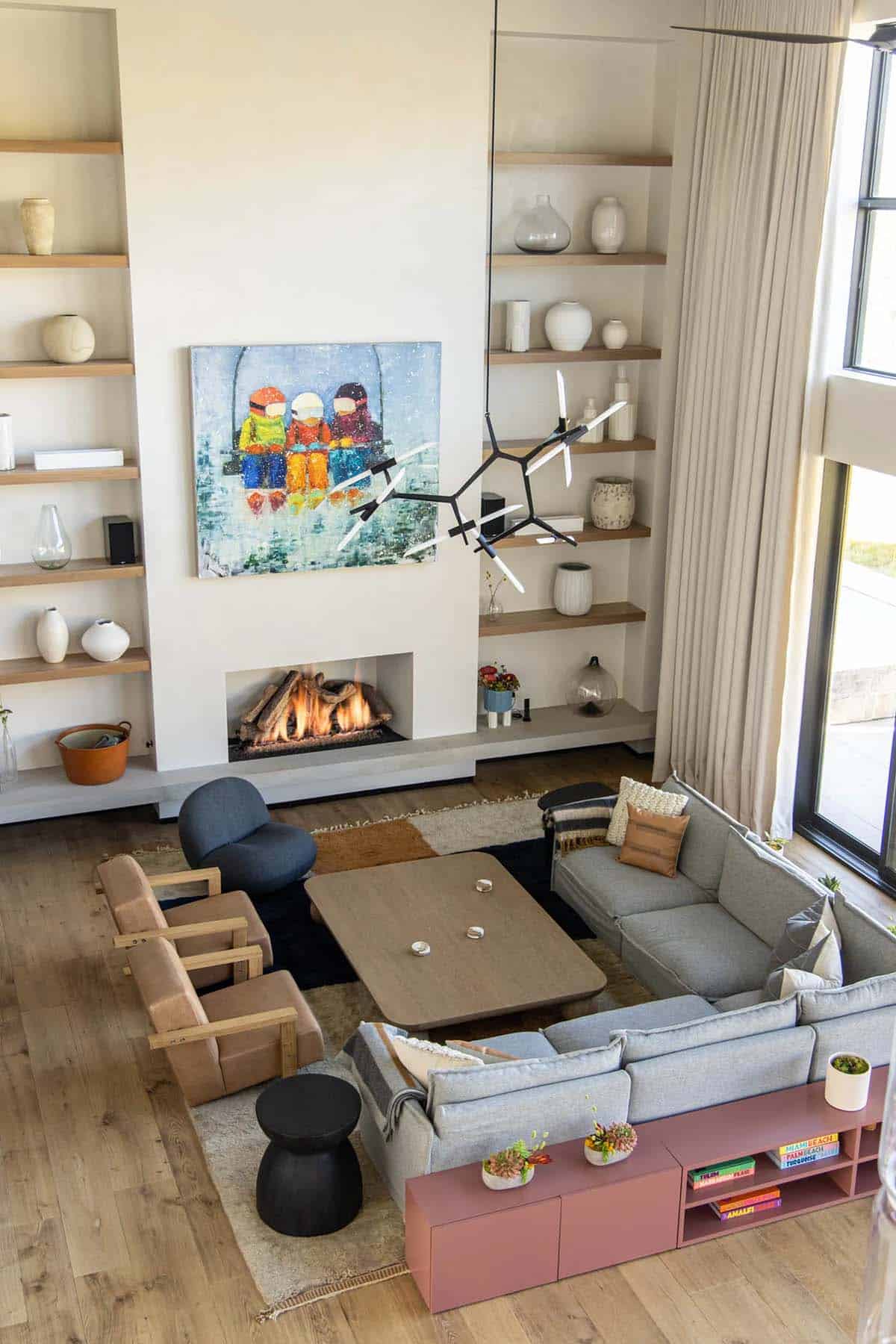Aerial view of living room with gray sectional, terracotta media console, lit fireplace, floor-to-ceiling shelves, and ski lift painting