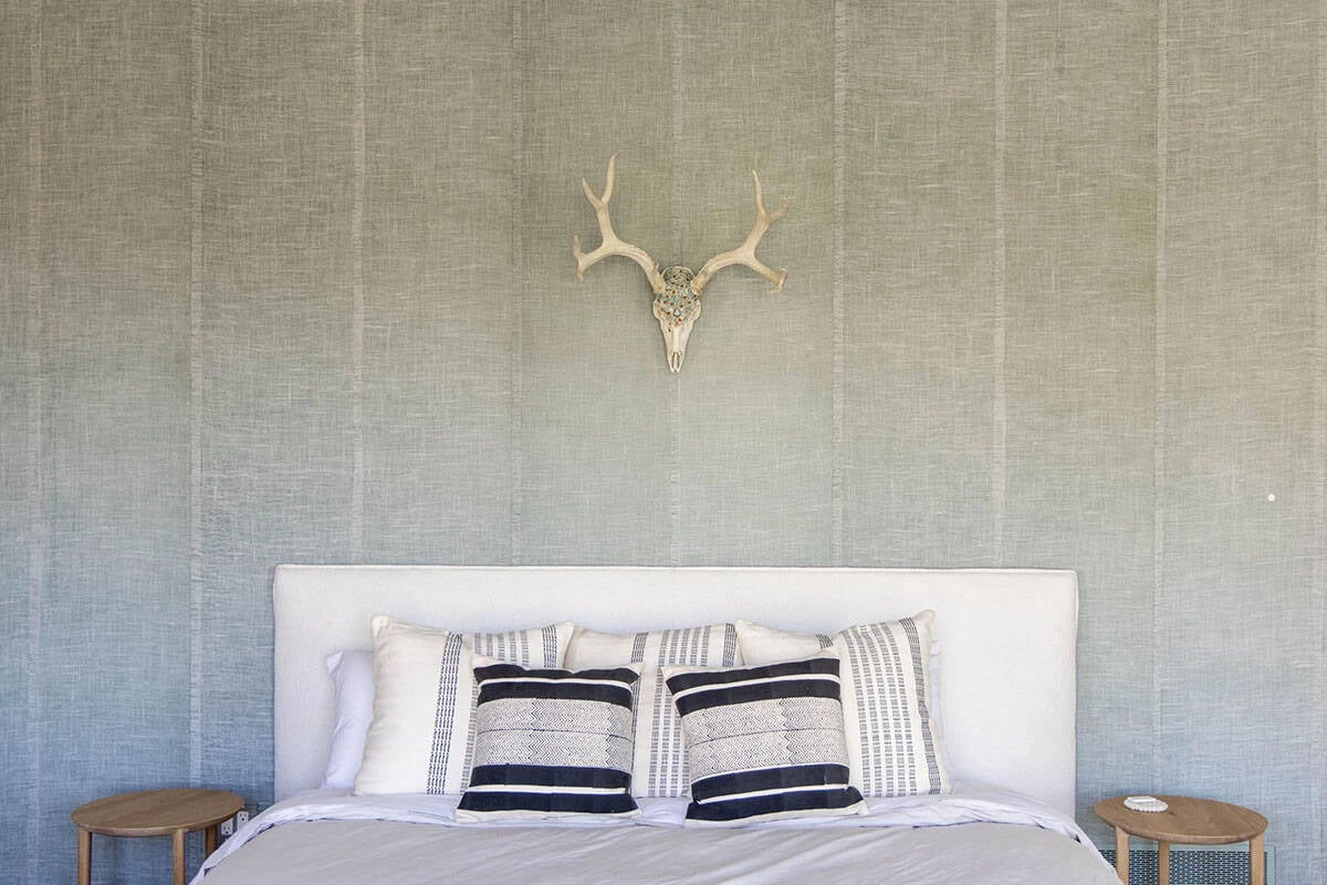 Bedroom headboard wall with sage green grasscloth wallcovering and decorative antler skull mount above white upholstered bed