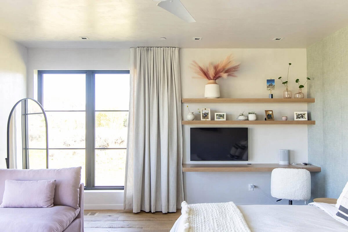Bedroom media nook with floating oak shelves styled with pampas grass, family photos, TV, and arched floor lamp beside linen drapes
